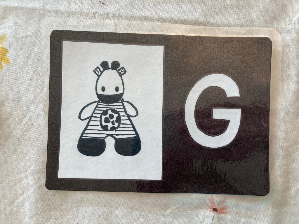 Photo of laminated card featuring black and white illustration of a toy giraffe and a cut paper letter G.
